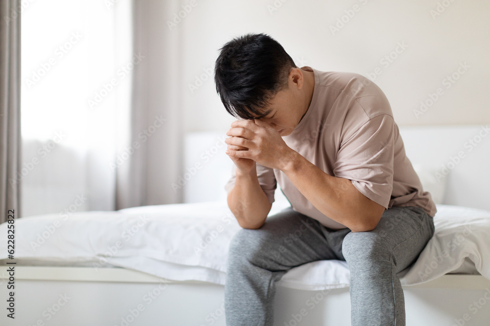 Unhappy middle aged asian man sitting on bed at home Unhappy middle aged asian man sitting on bed at home