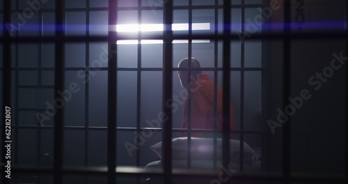 Elderly criminal in orange uniform sits in jail cell, stands up and looks on window with bars. Guilty inmate in detention center or correctional facility. Prisoner serves imprisonment term.