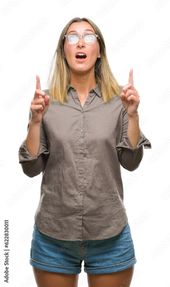 Young beautiful woman over isolated background amazed and surprised looking up and pointing with fingers and raised arms. Young beautiful woman over isolated background amazed and surprised looking up and pointing with fingers and raised arms.