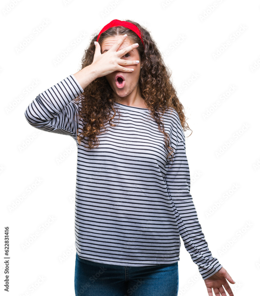 Beautiful brunette curly hair young girl wearing stripes sweater over isolated background peeking in shock covering face and eyes with hand, looking through fingers with embarrassed expression. Beautiful brunette curly hair young girl wearing stripes sweater over isolated background peeking in shock covering face and eyes with hand, looking through fingers with embarrassed expression.
