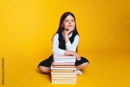Clever student girl wearing school uniform, holding hand under chin while sitting near pile of books, against yellow background Back to school concept