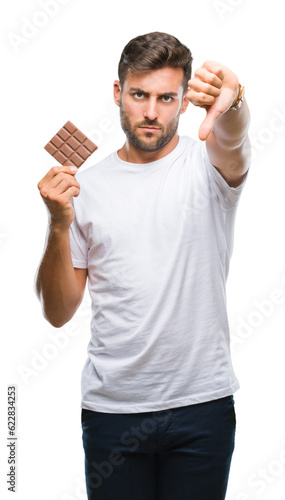 Young handsome man eating chocolate bar over isolated background with angry face, negative sign showing dislike with thumbs down, rejection concept
