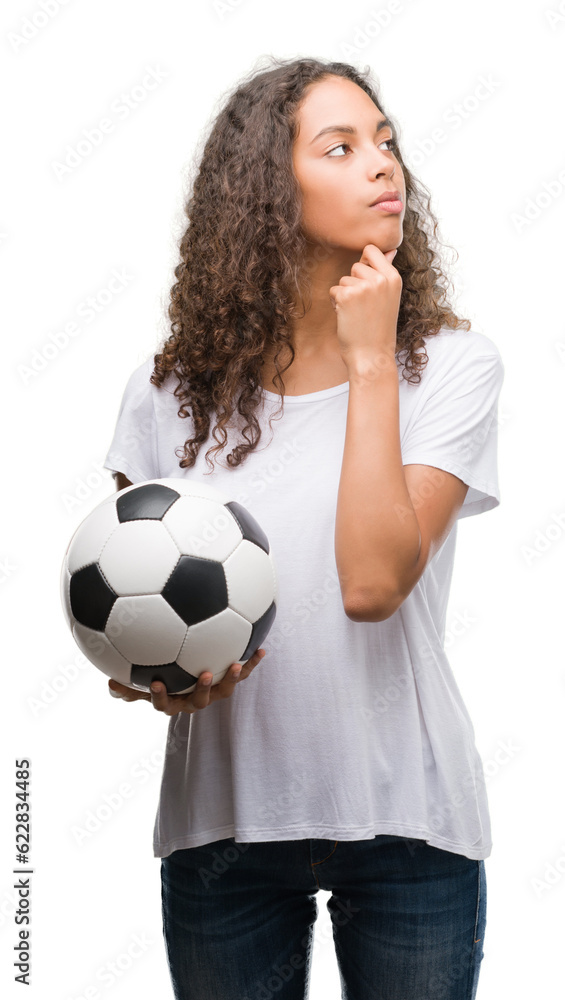 Young hispanic woman holding soccer football ball serious face thinking about question, very confused idea