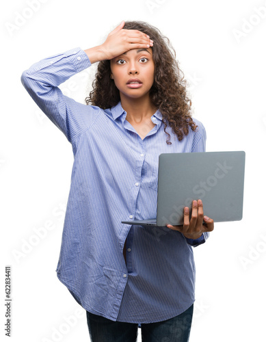 Young hispanic woman holding computer laptop stressed with hand on head, shocked with shame and surprise face, angry and frustrated. Fear and upset for mistake.