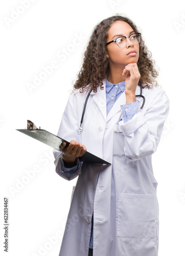 Фотография Young hispanic doctor woman holding a clipboard serious face thinking about ques
