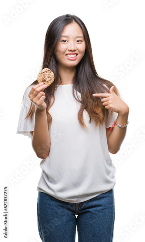 Young asian woman eating chocolate chip cookie over isolated background with surprise face pointing finger to himself