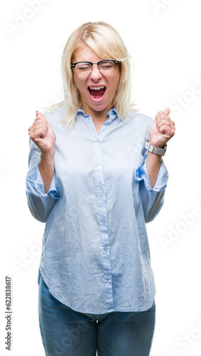 Young beautiful blonde business woman wearing glasses over isolated background excited for success with arms raised celebrating victory smiling. Winner concept.