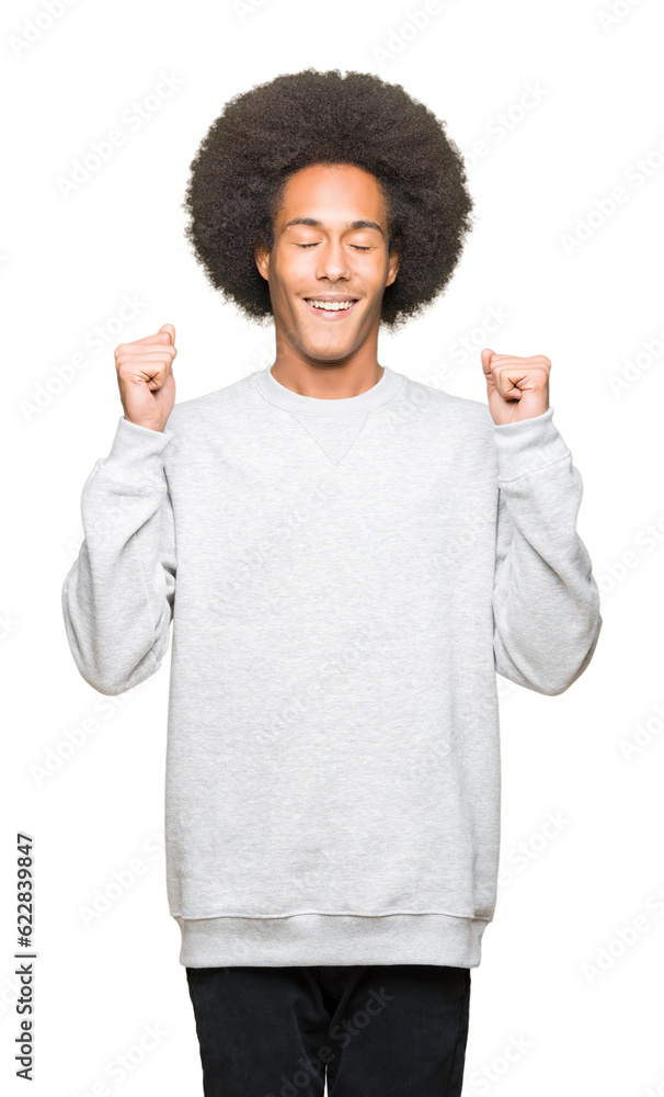 Young african american man with afro hair wearing sporty sweatshirt excited for success with arms raised celebrating victory smiling. Winner concept. Young african american man with afro hair wearing sporty sweatshirt excited for success with arms raised celebrating victory smiling. Winner concept.