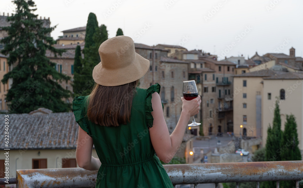Montepulciano, Province of Siena, Italy. Young woman in green dress drinking red wine and looking at ancient italian city at the evening