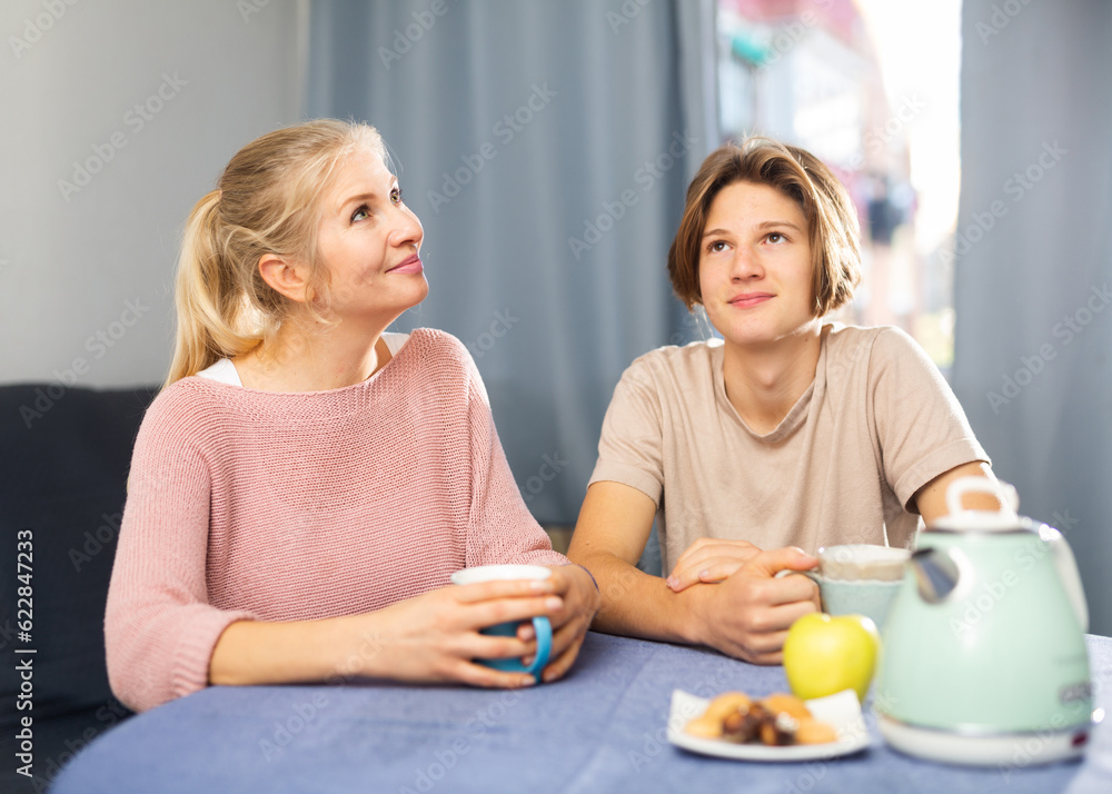 Smiling mother with teenage son drinking tea and talking at table at home indoors