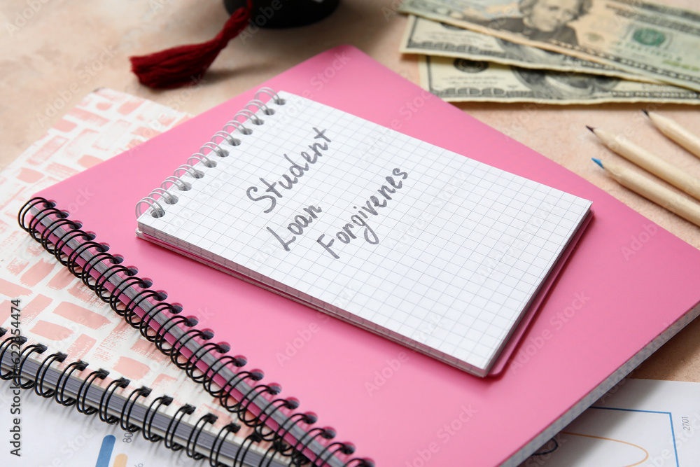 Notebook with text STUDENT LOAN FORGIVENESS, and dollar banknotes on beige table Notebook with text STUDENT LOAN FORGIVENESS, and dollar banknotes on beige table