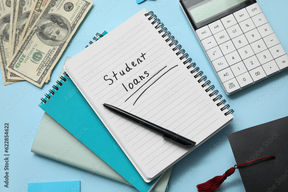 Notebook with text STUDENT LOANS, calculator, dollar banknotes and graduation cap on blue background Notebook with text STUDENT LOANS, calculator, dollar banknotes and graduation cap on blue background