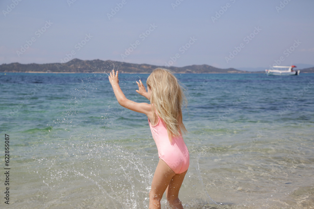 Naklejka premium Adorable little girl in pink swimsuit splashing water at the beach. Idyllic summer vacation concept.