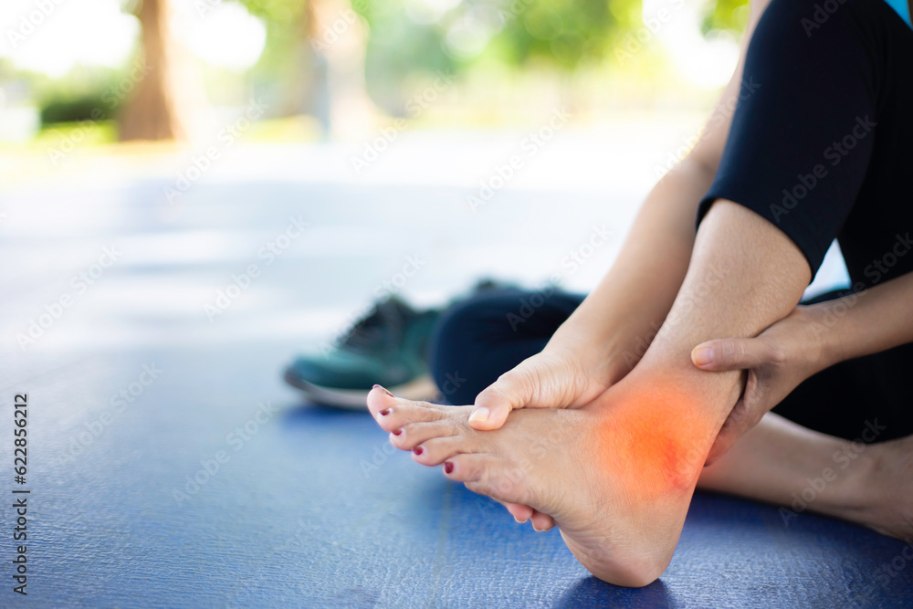Young women have pain in the ankle while exercising in a concrete path in the park. The blur bokeh background color of the leaves during the afternoons. Health care concept with exercise.