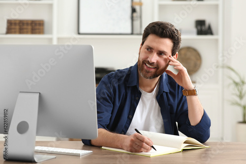 Home workplace. Happy man taking notes while working with computer at wooden desk in room