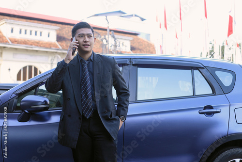 Young businessman in suit is standing near his car in front of modern business center and speaking on mobile phone