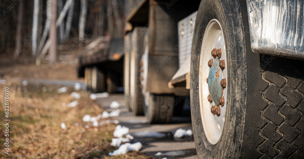 Front tire and wheel of a truck and trailer parked on a paved road on a sunny afternoon, with melting chunks of snow on the ground at job site. Shallow depth of field focus.