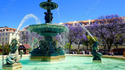 alter Brunnen mit Figuren und Wasserfontainen auf  Rossio Platz in Lissabon vor blauen Jacarandabäumen