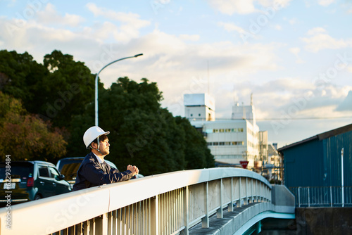 橋の上から遠くを見ている作業服とヘルメットの男性 photo