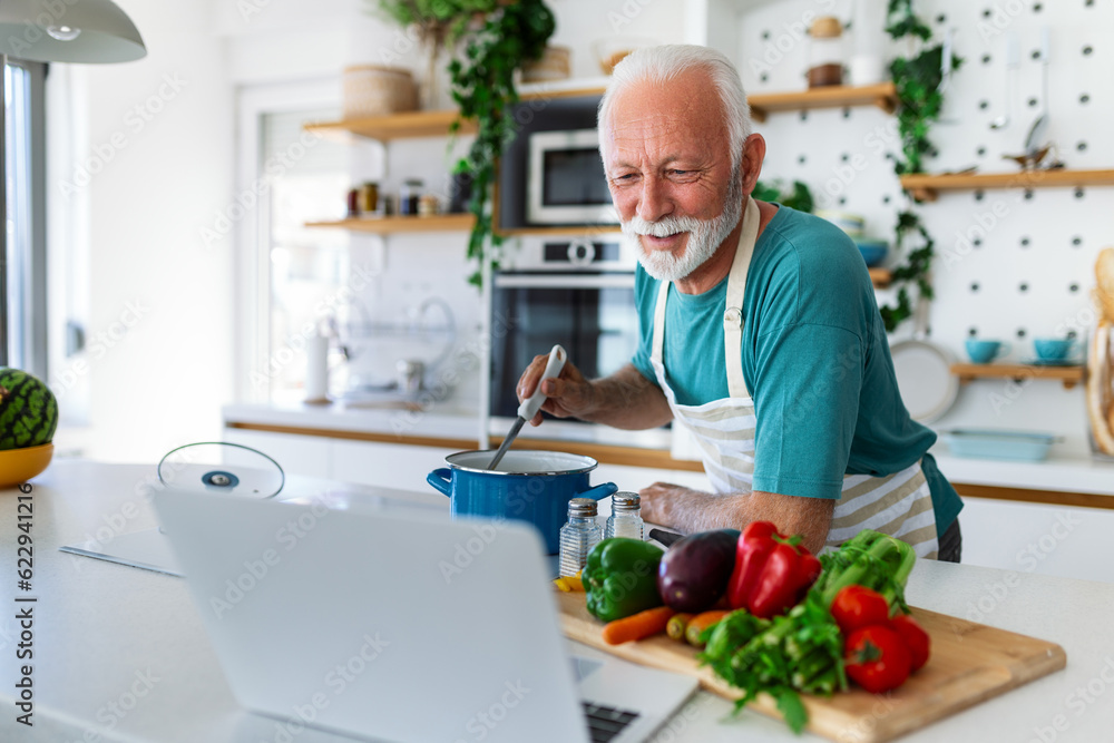 Happy senior man having fun cooking at home - Elderly person preparing healthy lunch in modern kitchen looking at the receipt at his laptop Happy senior man having fun cooking at home - Elderly person preparing healthy lunch in modern kitchen looking at the receipt at his laptop