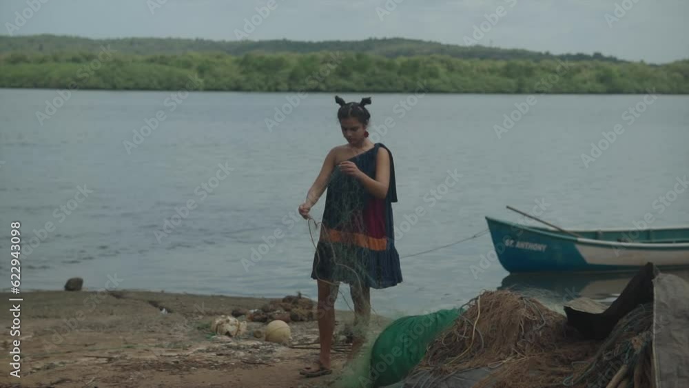 A young girl dressed in a navy elegant dress stands by the edge of a water reservoir, holding fishing nets in her hand. In the background, calm seawater and a few boats anchored to the shore. A young girl dressed in a navy elegant dress stands by the edge of a water reservoir, holding fishing nets in her hand. In the background, calm seawater and a few boats anchored to the shore.