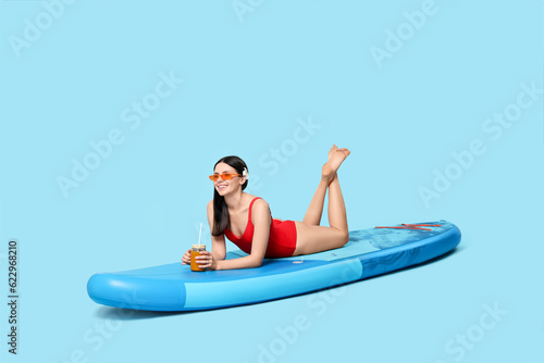 Happy woman with refreshing drink resting on SUP board against light blue background © New Africa