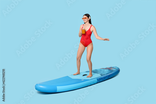 Happy woman with refreshing drink on SUP board against light blue background