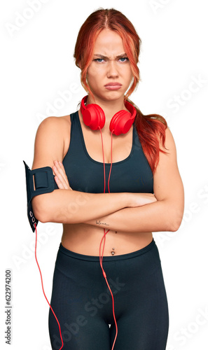 Young redhead woman wearing gym clothes and using headphones skeptic and nervous, disapproving expression on face with crossed arms. negative person.