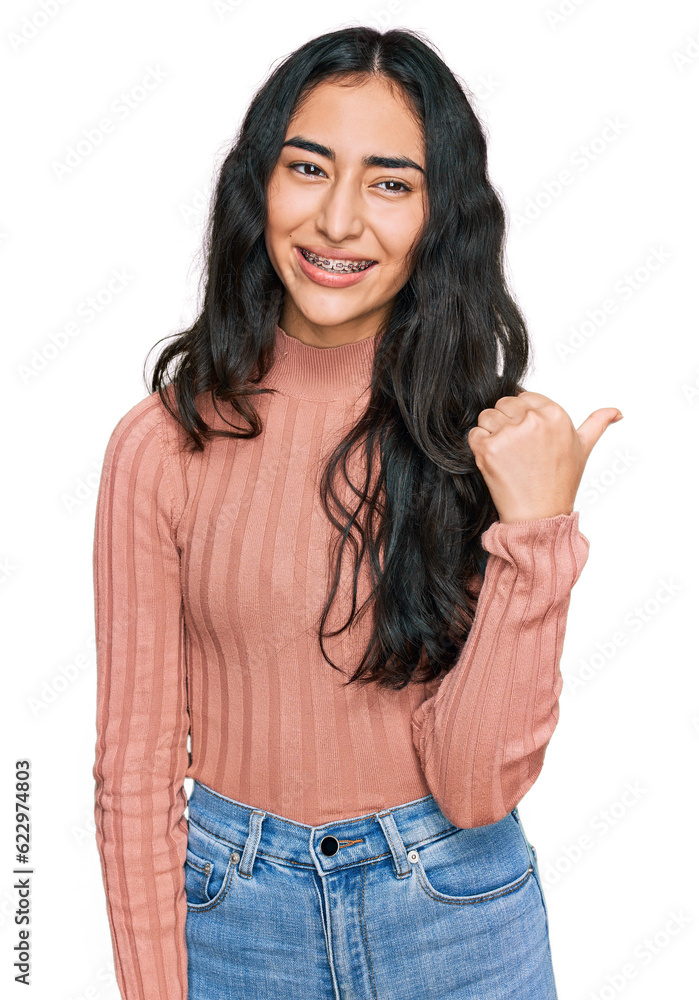 Hispanic teenager girl with dental braces wearing casual clothes smiling with happy face looking and pointing to the side with thumb up. Hispanic teenager girl with dental braces wearing casual clothes smiling with happy face looking and pointing to the side with thumb up.