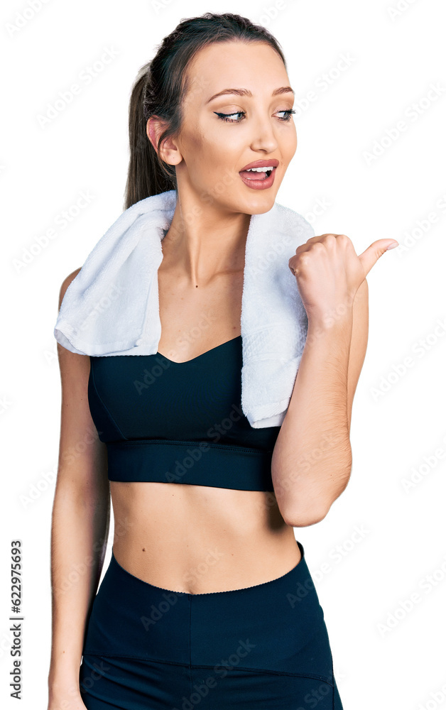 Beautiful caucasian woman wearing sportswear and towel smiling with happy face looking and pointing to the side with thumb up. Beautiful caucasian woman wearing sportswear and towel smiling with happy face looking and pointing to the side with thumb up.