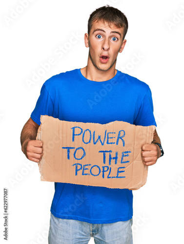 Young blond man holding power to the people banner scared and amazed with open mouth for surprise, disbelief face