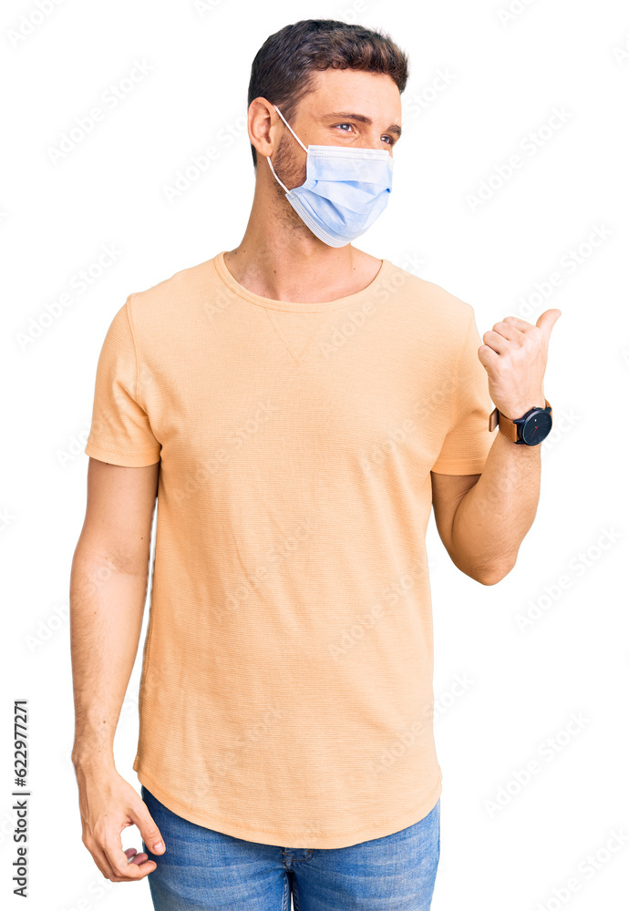 Handsome young man with bear wearing medical mask for coronavirus smiling with happy face looking and pointing to the side with thumb up.