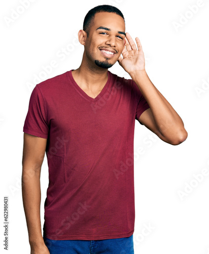 Young african american man wearing casual t shirt smiling with hand over ear listening and hearing to rumor or gossip. deafness concept.