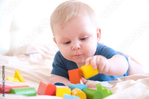 Tableau sur toile baby boy in blue bodysuit playing with colorful wooden eco toys on bed