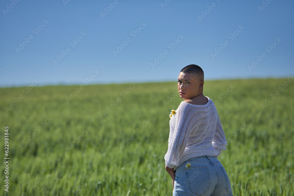 Young, South American, non-binary person, heavily makeup, posing in a white sweater with natural daisies, in the middle of a green wheat field. Concept queen, lgbtq+, pride, queer.