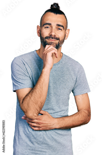 Hispanic man with ponytail wearing casual grey t shirt looking confident at the camera with smile with crossed arms and hand raised on chin. thinking positive.