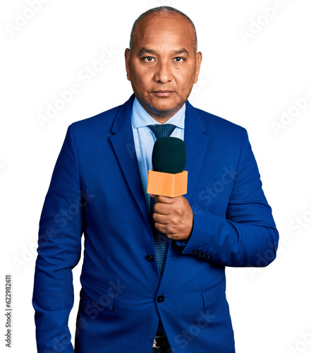 Hispanic middle age man holding reporter microphone thinking attitude and sober expression looking self confident