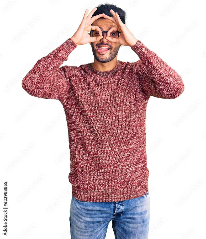 Young handsome hispanic man wearing winter sweater and glasses doing ok gesture like binoculars sticking tongue out, eyes looking through fingers. crazy expression. Young handsome hispanic man wearing winter sweater and glasses doing ok gesture like binoculars sticking tongue out, eyes looking through fingers. crazy expression.