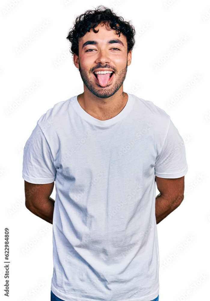Hispanic young man with beard wearing casual white t shirt sticking tongue out happy with funny expression. emotion concept. Hispanic young man with beard wearing casual white t shirt sticking tongue out happy with funny expression. emotion concept.