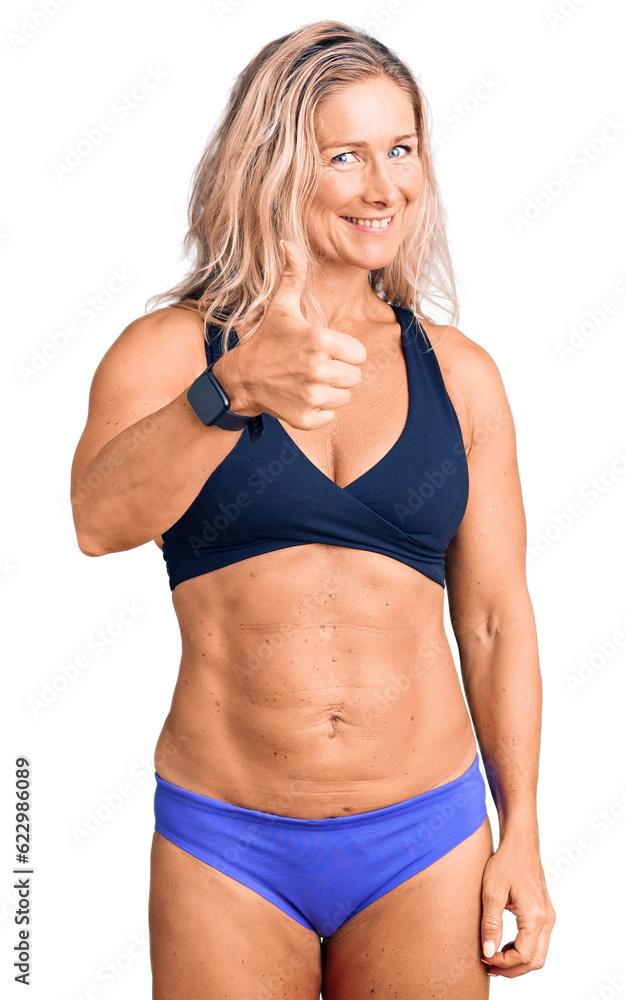 Middle age fit blonde woman wearing bikini doing happy thumbs up gesture with hand. approving expression looking at the camera showing success. Middle age fit blonde woman wearing bikini doing happy thumbs up gesture with hand. approving expression looking at the camera showing success.