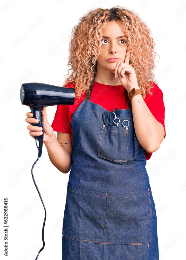 Young blonde woman with curly hair wearing hairdresser apron and holding dryer blow serious face thinking about question with hand on chin, thoughtful about confusing idea Young blonde woman with curly hair wearing hairdresser apron and holding dryer blow serious face thinking about question with hand on chin, thoughtful about confusing idea