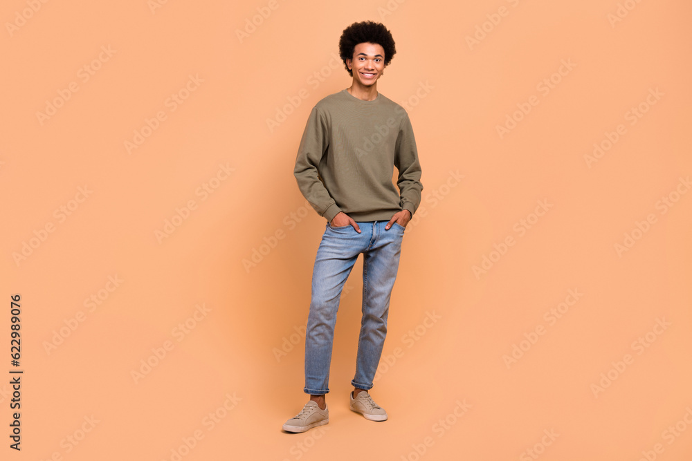 Full length photo of positive handsome guy wear stylish outfit hands pockets denim jeans brown sweatshirt model isolated on beige color background