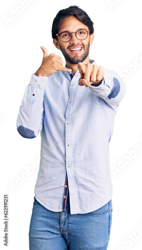 Handsome hispanic man wearing business shirt and glasses smiling doing talking on the telephone gesture and pointing to you. call me.