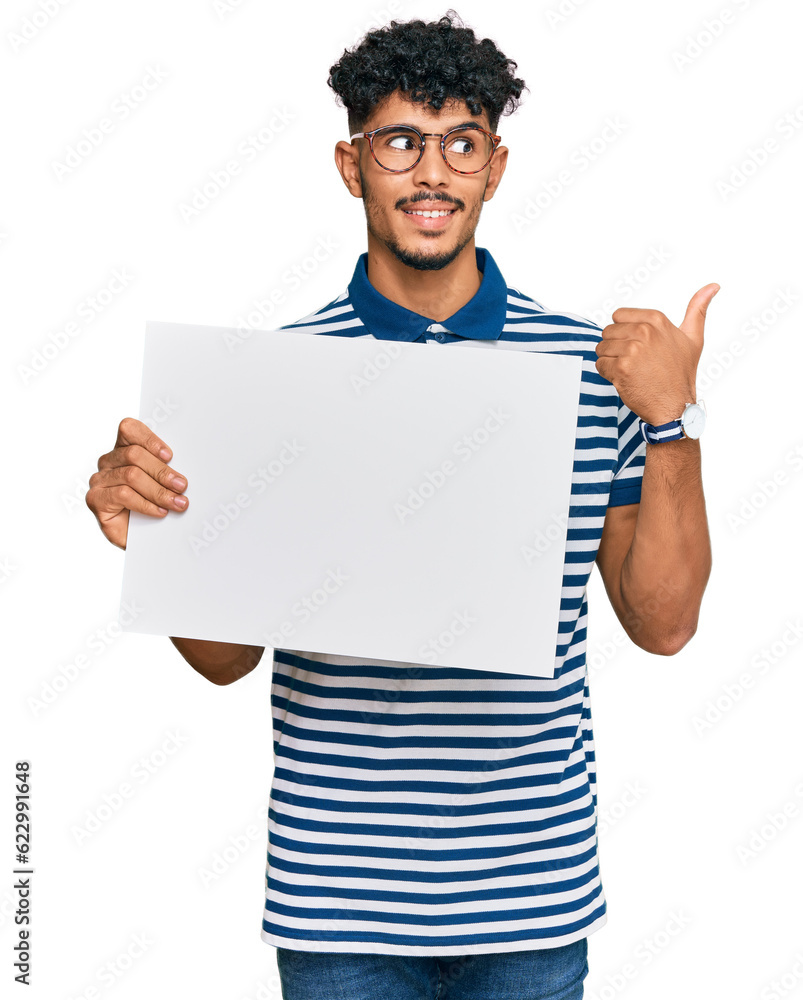 Young arab man holding blank empty banner pointing thumb up to the side smiling happy with open mouth Young arab man holding blank empty banner pointing thumb up to the side smiling happy with open mouth