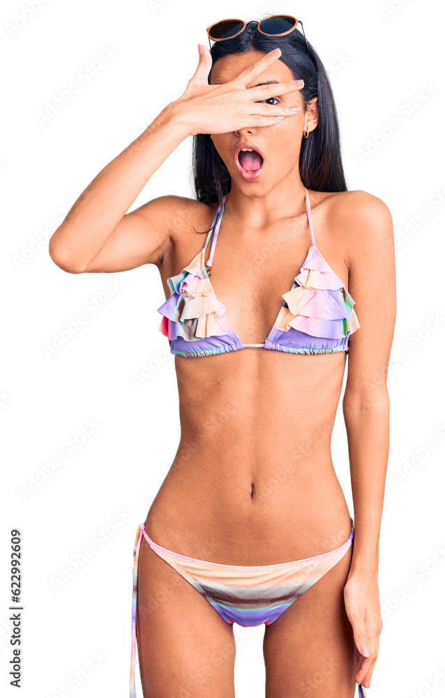Fototapeta premium Young beautiful latin girl wearing bikini and sunglasses peeking in shock covering face and eyes with hand, looking through fingers with embarrassed expression.