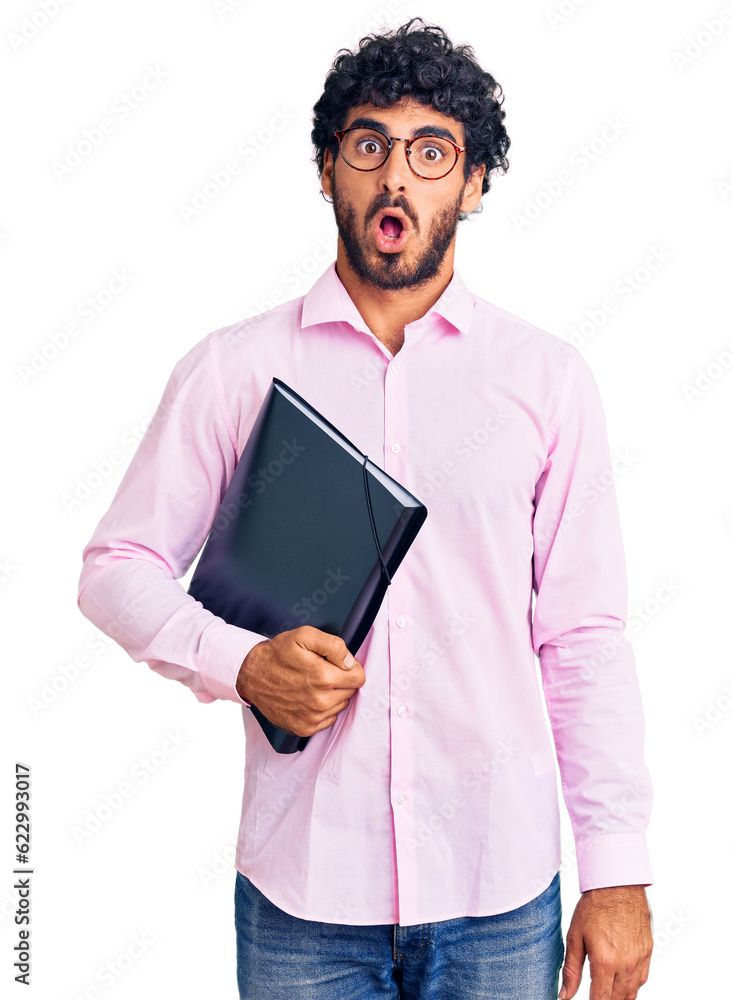 Handsome young man with curly hair and bear holding business folder scared and amazed with open mouth for surprise, disbelief face Handsome young man with curly hair and bear holding business folder scared and amazed with open mouth for surprise, disbelief face