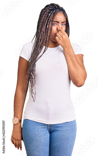 Young african american woman with braids wearing casual clothes smelling something stinky and disgusting, intolerable smell, holding breath with fingers on nose. bad smell © Krakenimages.com