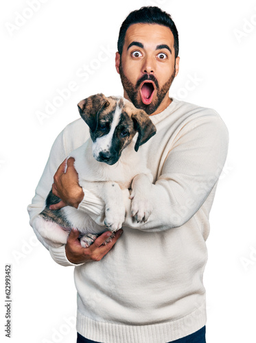 Young hispanic man with beard hugging cute dog afraid and shocked with surprise and amazed expression, fear and excited face.