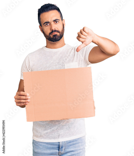 Young hispanic man holding banner cardboard with angry face, negative sign showing dislike with thumbs down, rejection concept