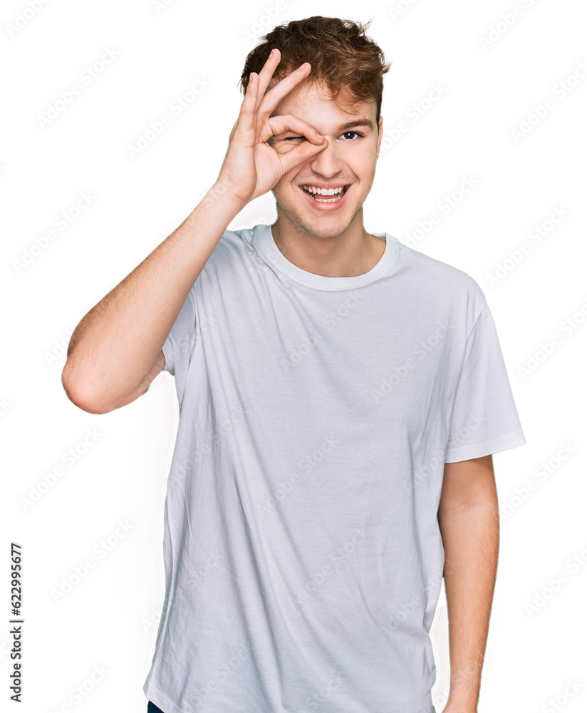 Young caucasian man wearing casual white t shirt doing ok gesture with hand smiling, eye looking through fingers with happy face. Young caucasian man wearing casual white t shirt doing ok gesture with hand smiling, eye looking through fingers with happy face.